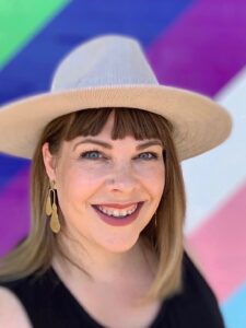 A photo of a white-presenting woman with a summer hat, and the inclusive pride flag colours in the background,